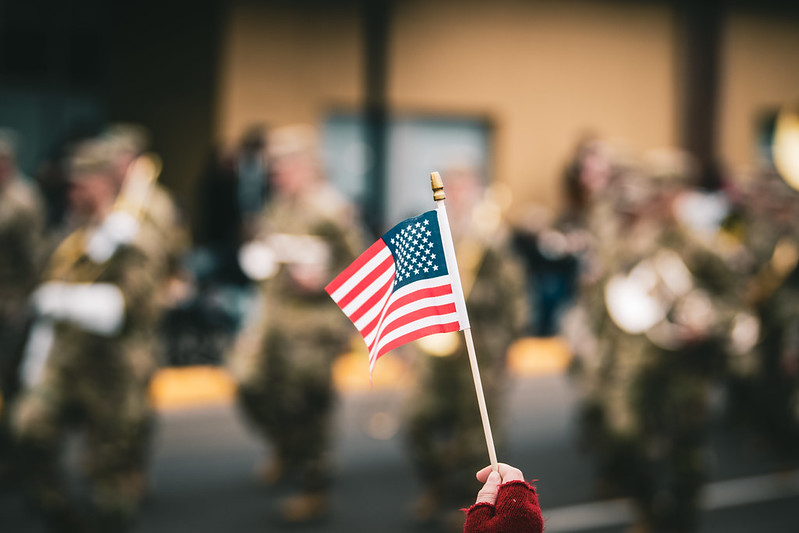 a hand holding a small American flag with a blurred background of soldiers